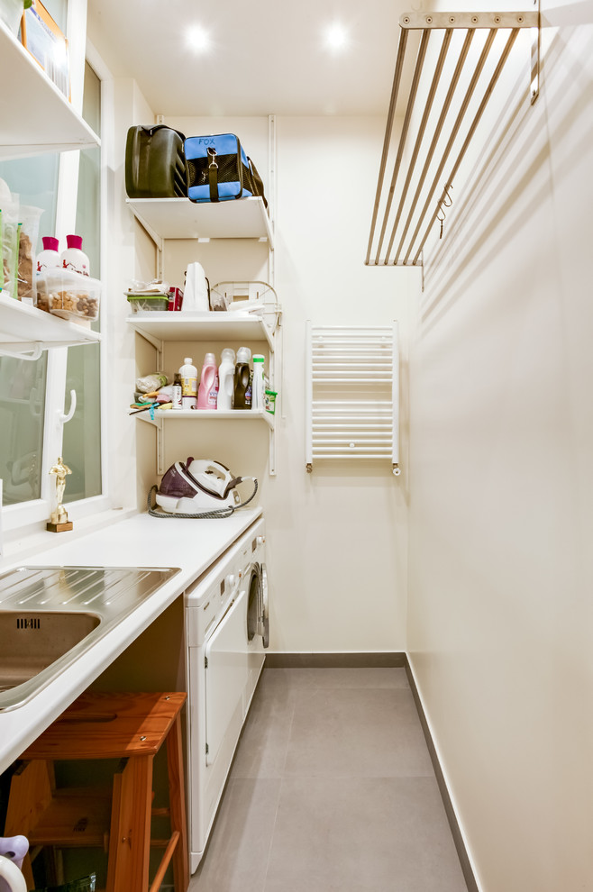 This is an example of a mid-sized modern single-wall utility room in Paris with an undermount sink, open cabinets, white walls, ceramic floors, a side-by-side washer and dryer and grey floor.