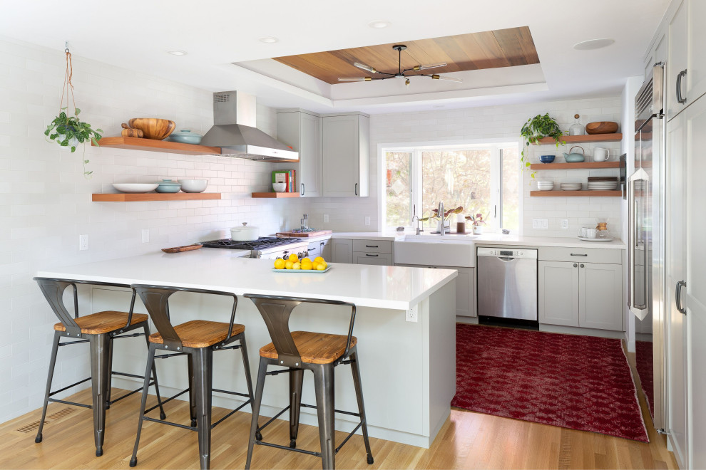 Example of a mid-sized transitional u-shaped light wood floor, brown floor and tray ceiling open concept kitchen design with a farmhouse sink, shaker cabinets, gray cabinets, quartz countertops, white backsplash, ceramic backsplash, stainless steel appliances, a peninsula and white countertops