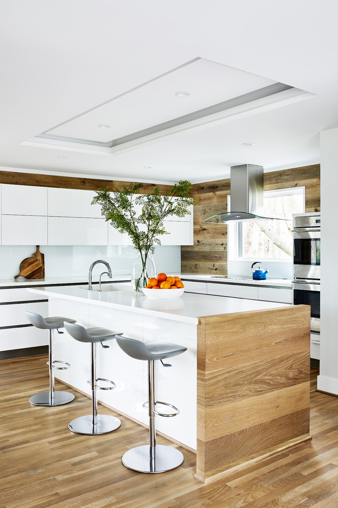 Photo of a mid-sized midcentury l-shaped eat-in kitchen in DC Metro with flat-panel cabinets, white cabinets, solid surface benchtops, white splashback, stainless steel appliances, light hardwood floors, with island, beige floor, white benchtop, an integrated sink and glass sheet splashback.
