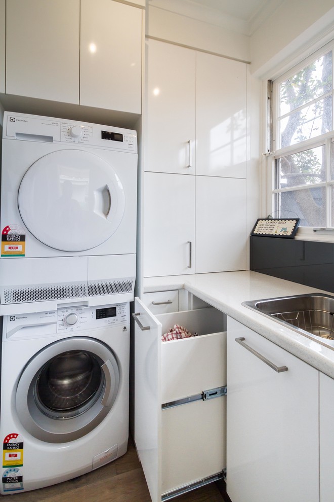 Design ideas for a small modern l-shaped utility room in Melbourne with flat-panel cabinets, white cabinets, laminate benchtops, white walls, light hardwood floors, a stacked washer and dryer and a drop-in sink.