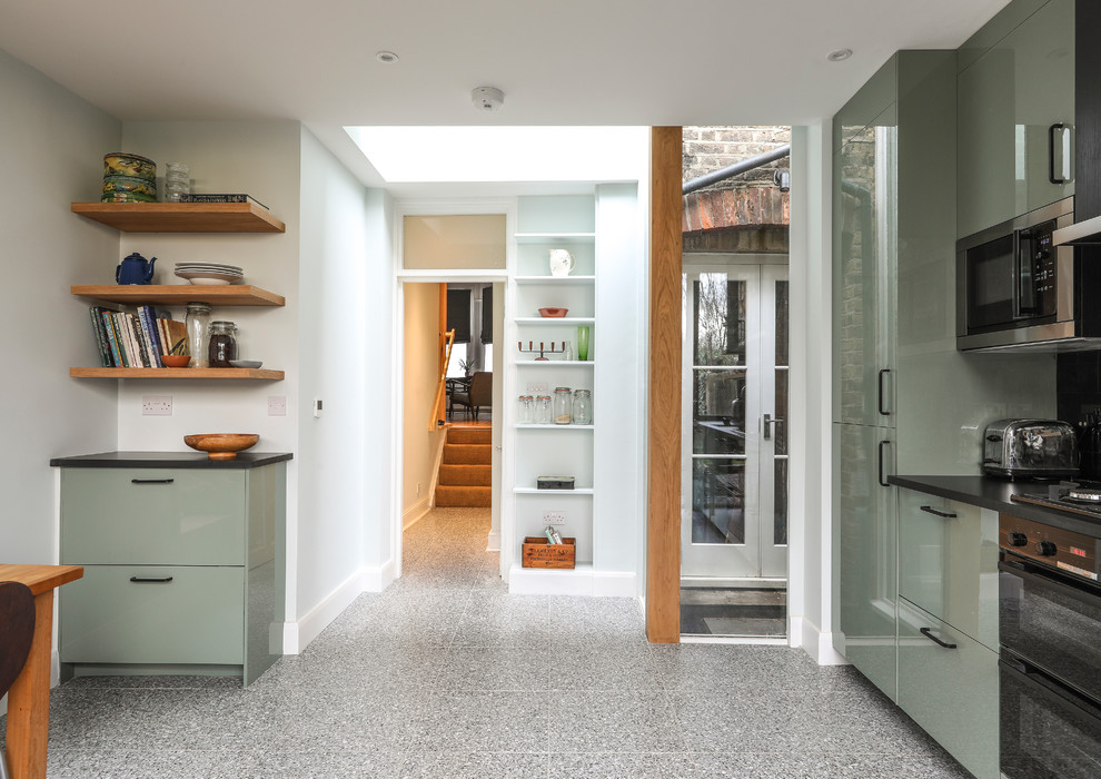 Photo of a mid-sized contemporary single-wall open plan kitchen in London with a drop-in sink, flat-panel cabinets, green cabinets, solid surface benchtops, black splashback, ceramic splashback, stainless steel appliances, terrazzo floors, grey floor and black benchtop.