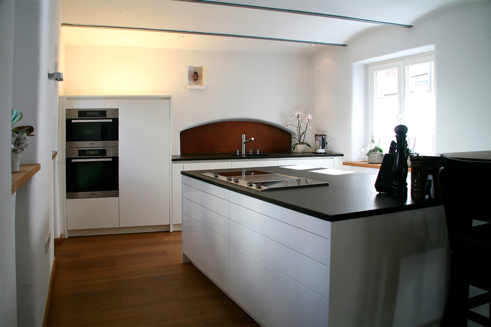 Küche in L-Form With Unterbauwaschbecken, flächenbündigen Schrankfronten, weißen Schränken, Granit-Arbeitsplatte, Küchenrückwand in Braun, hellem Holzboden, Halbinsel, beigem Boden and schwarzer Arbeitsplatte in Nuremberg