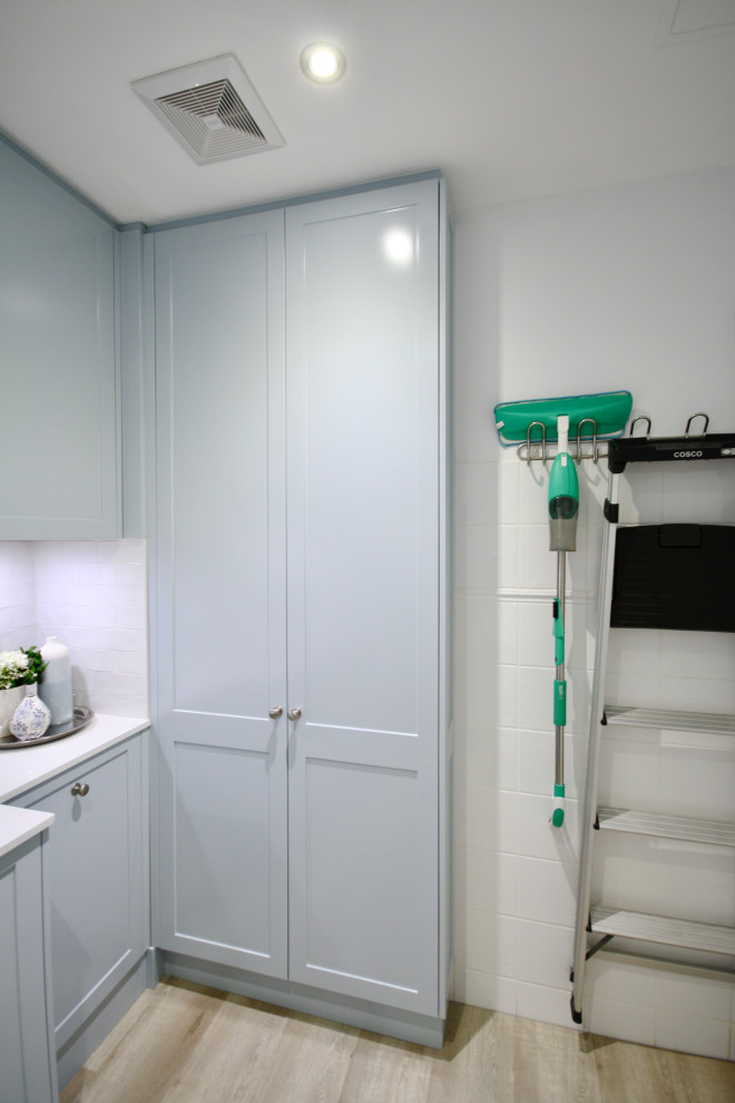 This is an example of a mid-sized transitional u-shaped dedicated laundry room in Sydney with an undermount sink, shaker cabinets, blue cabinets, quartz benchtops, white splashback, subway tile splashback, white walls, light hardwood floors, a stacked washer and dryer, brown floor and white benchtop.