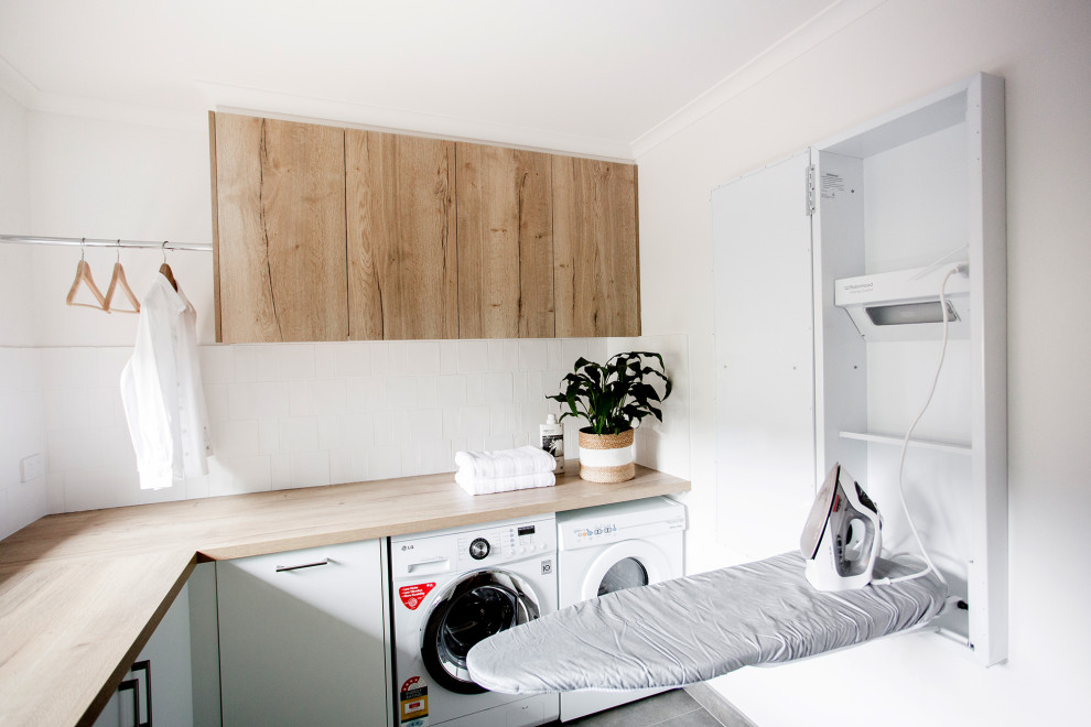 This is an example of a mid-sized contemporary l-shaped dedicated laundry room in Other with a drop-in sink, flat-panel cabinets, medium wood cabinets, wood benchtops, white splashback, ceramic splashback, white walls, ceramic floors, a side-by-side washer and dryer and grey floor.