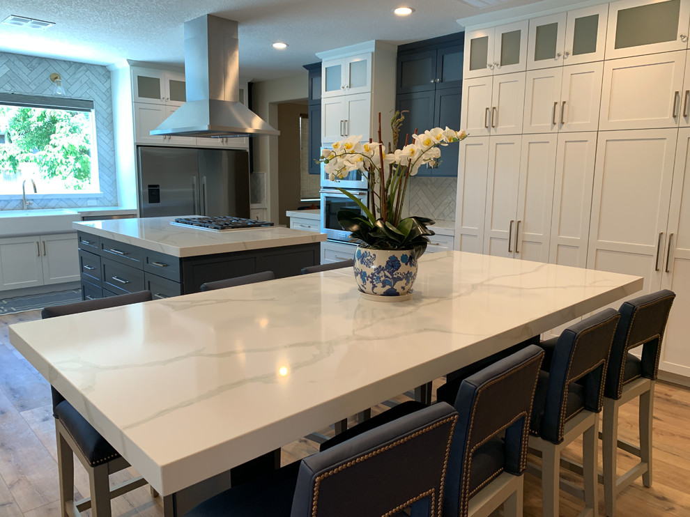 Large transitional l-shaped open plan kitchen in Sacramento with a farmhouse sink, shaker cabinets, white cabinets, marble benchtops, grey splashback, subway tile splashback, stainless steel appliances, light hardwood floors, with island, beige floor and white benchtop.