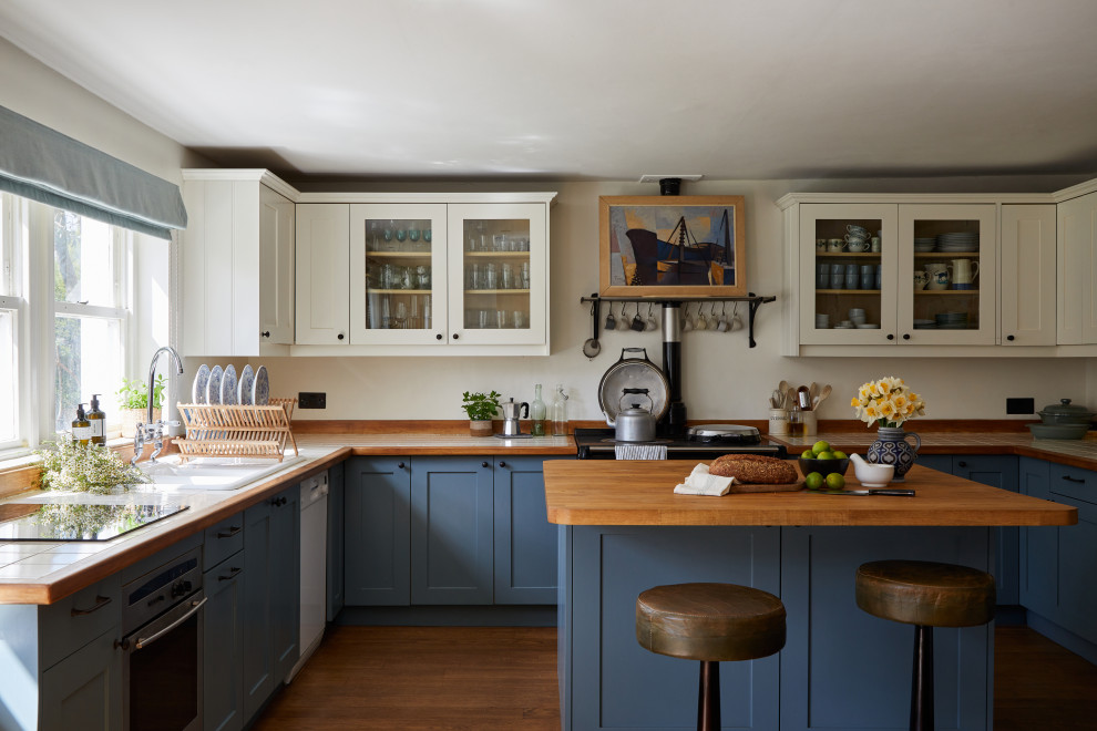 Photo of a traditional u-shaped kitchen/diner in London with a double-bowl sink, shaker cabinets, blue cabinets, wood worktops, wood splashback, medium hardwood flooring, an island and brown floors.