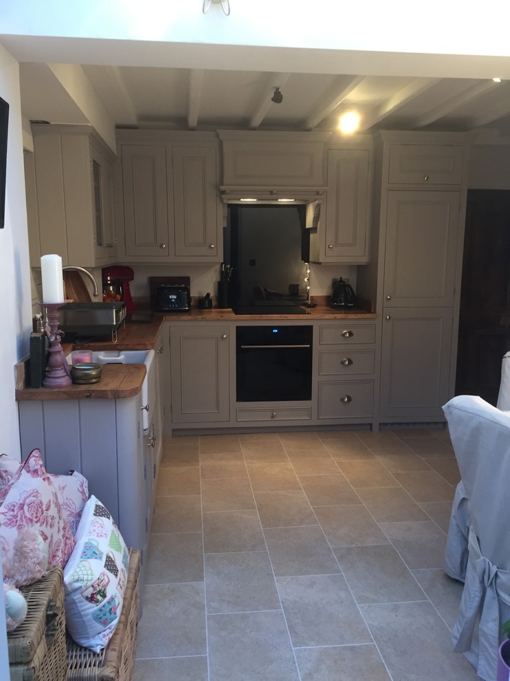 Photo of a small traditional l-shaped eat-in kitchen in Other with a farmhouse sink, shaker cabinets, grey cabinets, wood benchtops, black splashback, glass sheet splashback, black appliances, laminate floors and no island.