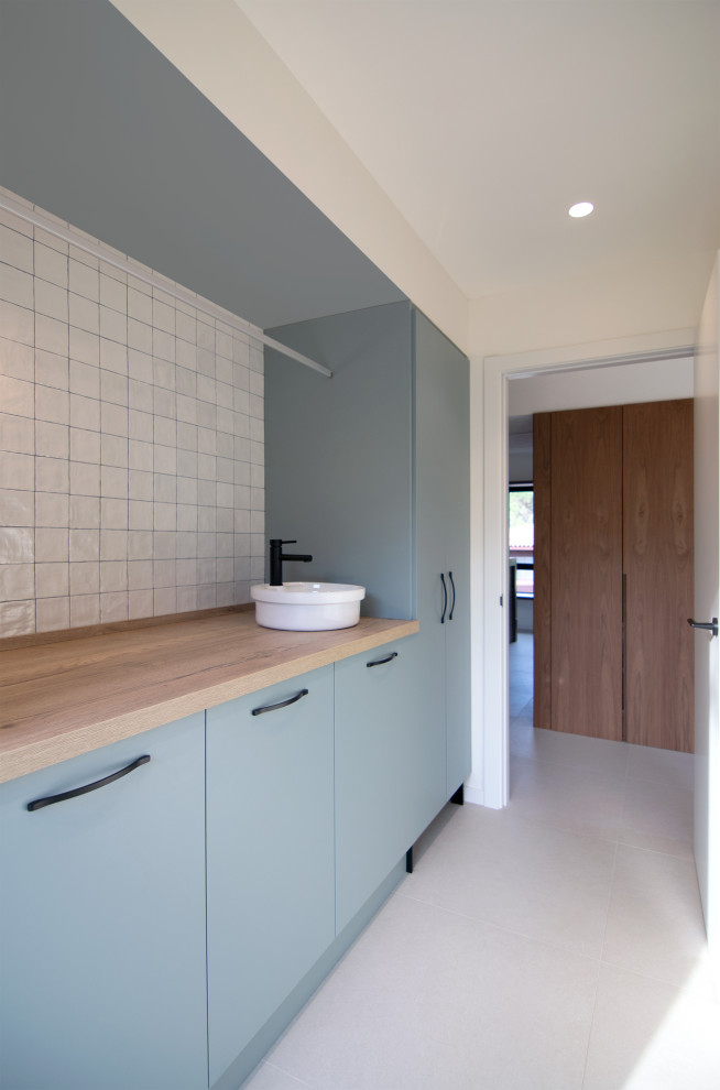 Example of a mid-sized trendy single-wall ceramic tile and beige floor dedicated laundry room design in Barcelona with a farmhouse sink, flat-panel cabinets, turquoise cabinets, wood countertops, white backsplash, ceramic backsplash, white walls, a concealed washer/dryer and brown countertops