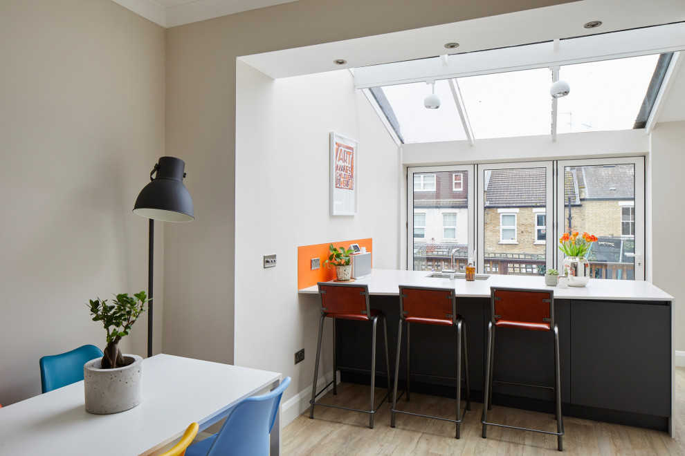 Medium sized contemporary kitchen/diner in London with a submerged sink, flat-panel cabinets, white cabinets, engineered stone countertops, orange splashback, glass sheet splashback, stainless steel appliances, light hardwood flooring, an island and white worktops.