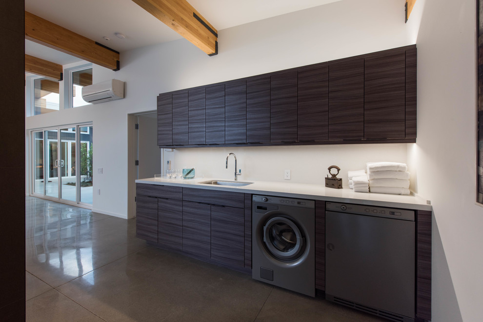 Photo of a mid-sized midcentury single-wall utility room in Other with a single-bowl sink, flat-panel cabinets, dark wood cabinets, solid surface benchtops, white walls, concrete floors, a side-by-side washer and dryer and grey floor.