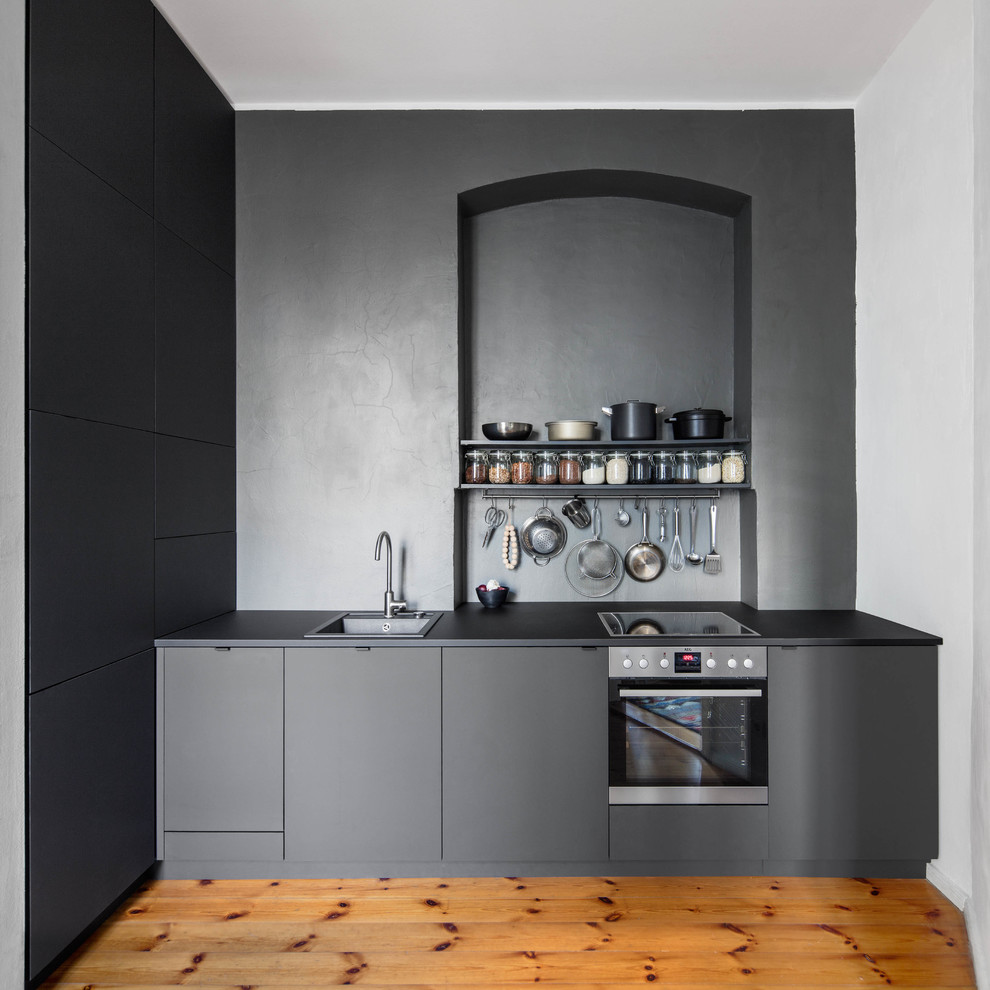 Mid-sized modern single-wall open plan kitchen in Berlin with a drop-in sink, flat-panel cabinets, black cabinets, solid surface benchtops, stone slab splashback, no island, brown floor, black splashback, medium hardwood floors, black benchtop and stainless steel appliances.