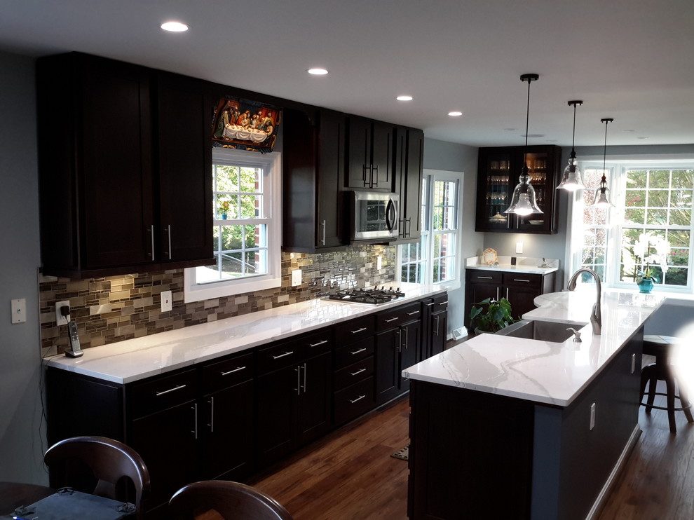 Large traditional u-shaped eat-in kitchen in DC Metro with a farmhouse sink, shaker cabinets, dark wood cabinets, quartz benchtops, brown splashback, glass tile splashback, stainless steel appliances, medium hardwood floors, with island, brown floor and white benchtop.