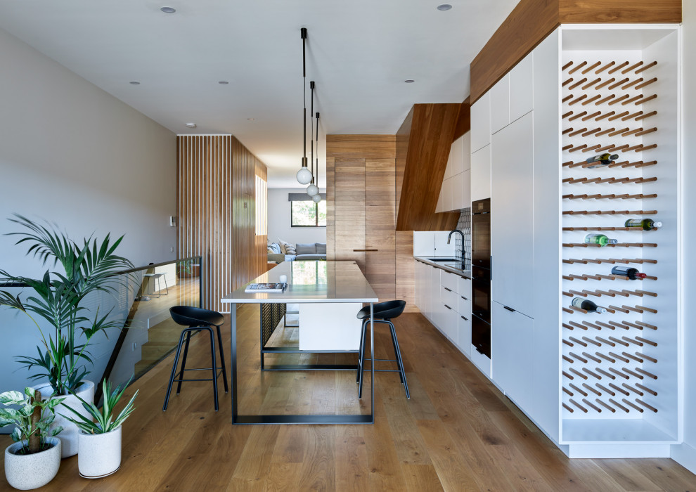 Design ideas for a large contemporary galley open plan kitchen in Melbourne with an undermount sink, white cabinets, quartz benchtops, white splashback, ceramic splashback, black appliances, light hardwood floors, with island, brown floor and grey benchtop.