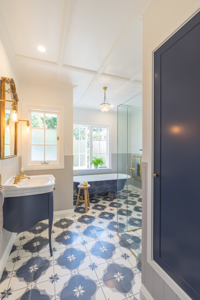 This is an example of a mid-sized transitional kids bathroom in Auckland with furniture-like cabinets, blue cabinets, a claw-foot tub, a curbless shower, a two-piece toilet, gray tile, ceramic tile, white walls, cement tiles, a console sink, blue floor and a hinged shower door.