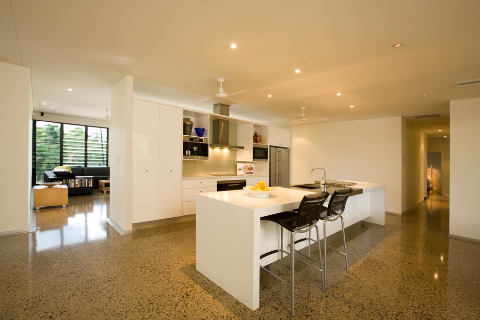 This is an example of a tropical galley open plan kitchen in Darwin with an integrated sink, flat-panel cabinets, white cabinets, solid surface benchtops, metallic splashback, glass sheet splashback, stainless steel appliances, concrete floors and with island.