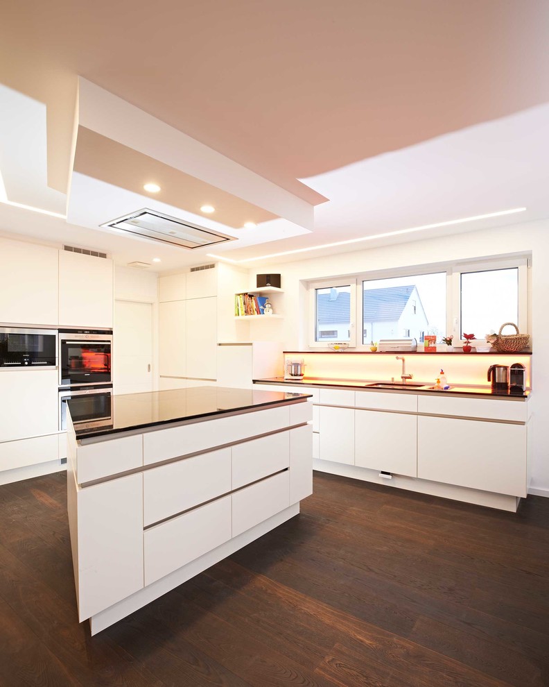 Photo of a mid-sized contemporary l-shaped open plan kitchen in Stuttgart with flat-panel cabinets, white cabinets, white splashback, glass sheet splashback, black appliances, dark hardwood floors, with island, brown floor and black benchtop.