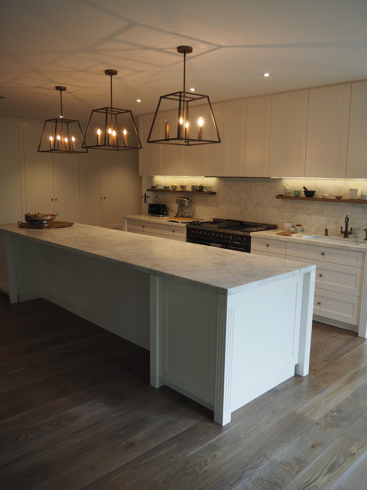 Photo of an expansive transitional galley open plan kitchen in Melbourne with a farmhouse sink, recessed-panel cabinets, white cabinets, marble benchtops, white splashback, marble splashback, black appliances, light hardwood floors, with island and brown floor.