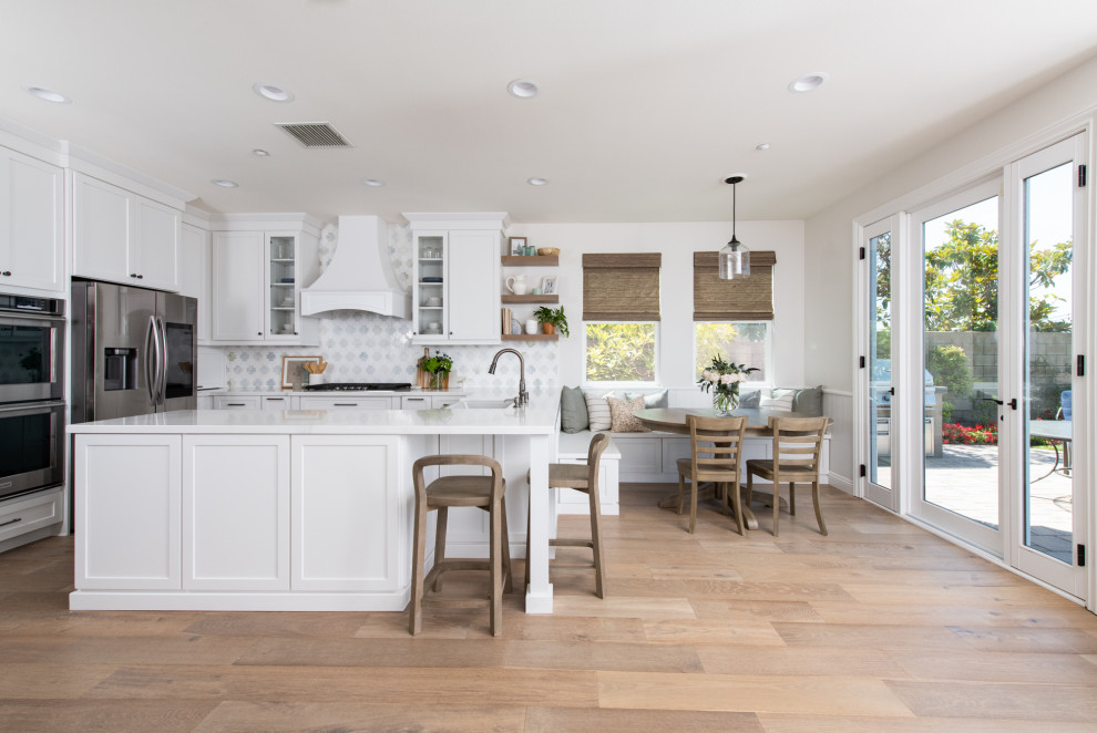 Design ideas for a mid-sized beach style l-shaped eat-in kitchen in Orange County with a farmhouse sink, recessed-panel cabinets, white cabinets, quartz benchtops, multi-coloured splashback, marble splashback, medium hardwood floors, with island, brown floor and white benchtop.