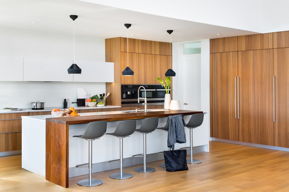 Modern kitchen in Vancouver with an undermount sink, flat-panel cabinets, medium wood cabinets, white splashback, black appliances, medium hardwood floors, with island and white benchtop.