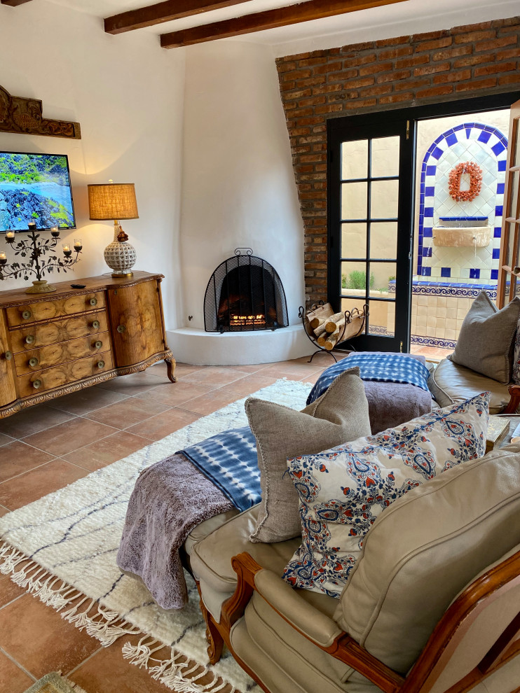 Mid-sized mediterranean open concept living room in San Diego with white walls, porcelain floors, a corner fireplace, a plaster fireplace surround, a wall-mounted tv, orange floor, exposed beam, a library and brick walls.