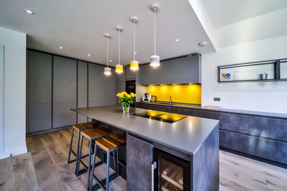 Photo of a large modern l-shaped open plan kitchen in London with flat-panel cabinets, grey cabinets, engineered stone countertops, yellow splashback, black appliances, an island and grey worktops.