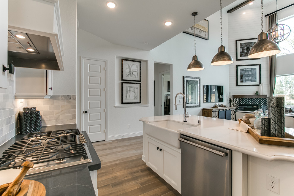 Photo of a modern galley open plan kitchen in Houston with a farmhouse sink, shaker cabinets, white cabinets, quartzite benchtops, grey splashback, stone tile splashback, stainless steel appliances, dark hardwood floors, with island, brown floor and grey benchtop.