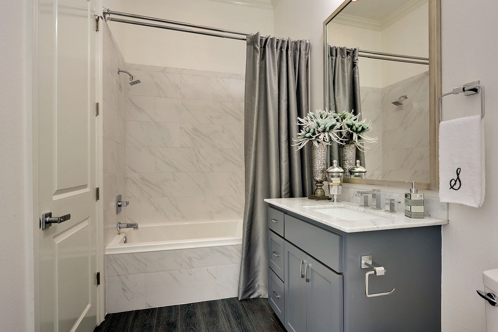 This is an example of a small modern 3/4 bathroom in New Orleans with shaker cabinets, grey cabinets, a shower/bathtub combo, a two-piece toilet, white tile, marble, beige walls, dark hardwood floors, an undermount sink, marble benchtops, brown floor and a shower curtain.