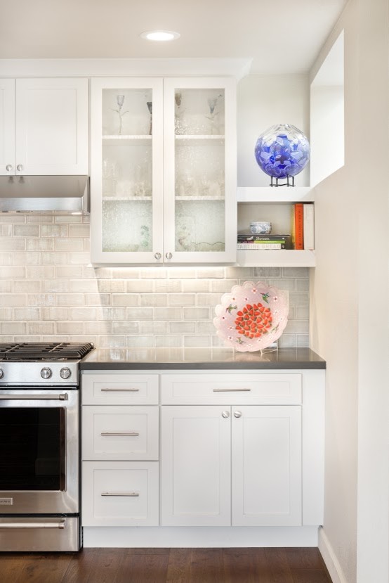 Example of a mid-sized transitional l-shaped medium tone wood floor eat-in kitchen design with glass-front cabinets, white cabinets, quartz countertops, gray backsplash, ceramic backsplash and stainless steel appliances