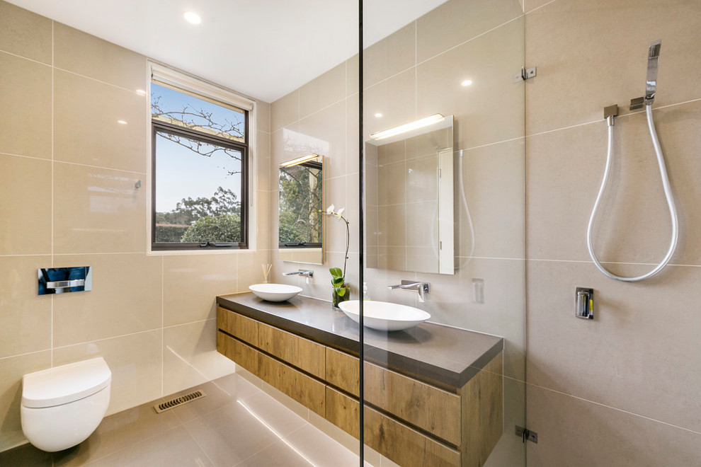 Photo of a contemporary bathroom in Melbourne with medium wood cabinets, a freestanding tub, an open shower, a wall-mount toilet, beige tile, porcelain tile, beige walls, porcelain floors, a vessel sink, engineered quartz benchtops, beige floor, an open shower, brown benchtops and flat-panel cabinets.