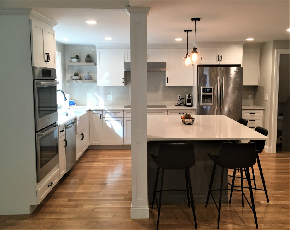 Example of a large transitional u-shaped light wood floor and brown floor eat-in kitchen design in Boston with a farmhouse sink, shaker cabinets, white cabinets, quartz countertops, beige backsplash, subway tile backsplash, stainless steel appliances, an island and white countertops