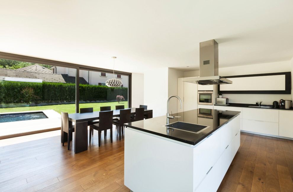 Large contemporary eat-in kitchen in Essen with white cabinets, a drop-in sink, flat-panel cabinets, medium hardwood floors, with island, white splashback and stainless steel appliances.