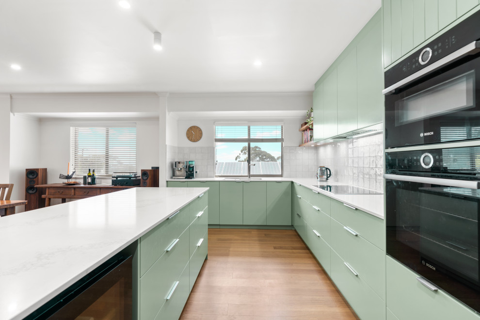 This is an example of a mid-sized contemporary l-shaped kitchen in Adelaide with a drop-in sink, green cabinets, quartz benchtops, white splashback, porcelain splashback, black appliances, medium hardwood floors, with island and white benchtop.