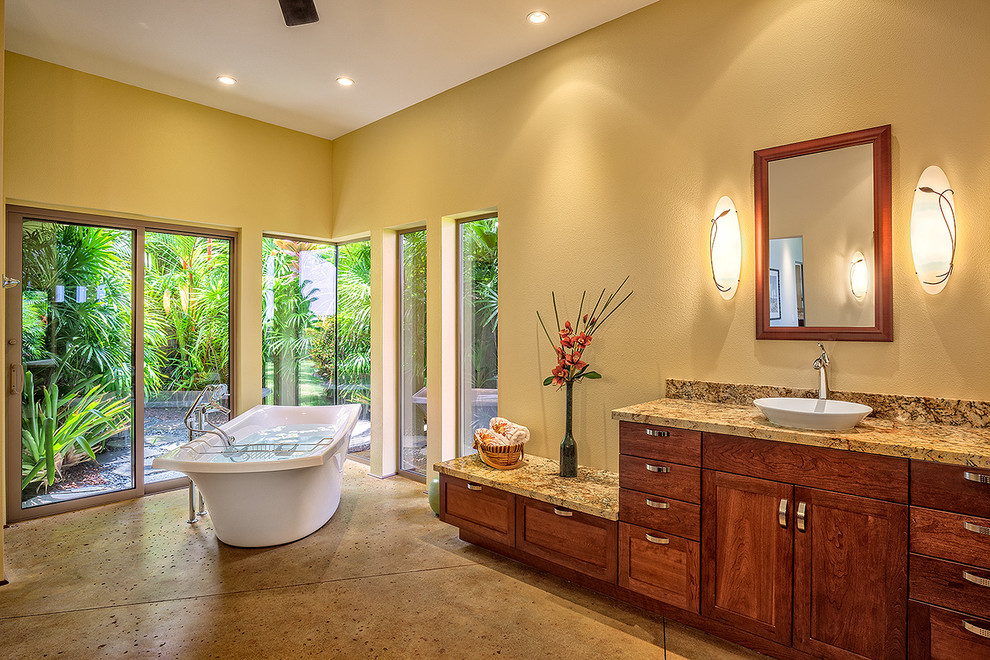Expansive traditional master bathroom in Hawaii with a drop-in sink, recessed-panel cabinets, medium wood cabinets, granite benchtops, a freestanding tub, a double shower, beige tile, stone slab, beige walls, concrete floors and beige floor.