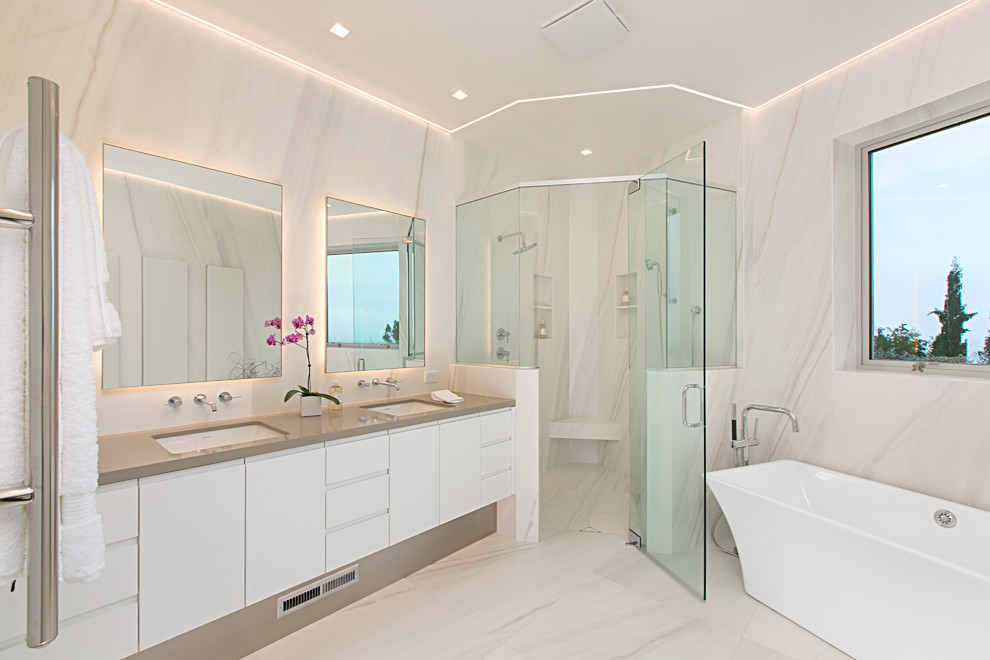 This is an example of a contemporary master bathroom in San Diego with flat-panel cabinets, white cabinets, a freestanding tub, a corner shower, white tile, marble, white walls, marble floors, an undermount sink, white floor, a hinged shower door and grey benchtops.