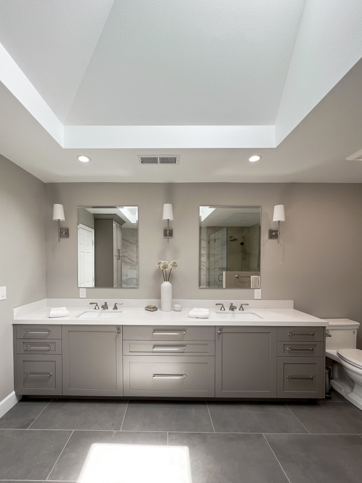 Photo of a mid-sized transitional master bathroom in San Francisco with shaker cabinets, brown cabinets, a built-in vanity, a freestanding tub, a corner shower, a one-piece toilet, white tile, porcelain tile, white walls, porcelain floors, an undermount sink, engineered quartz benchtops, grey floor, a hinged shower door, white benchtops, a niche, a double vanity and recessed.