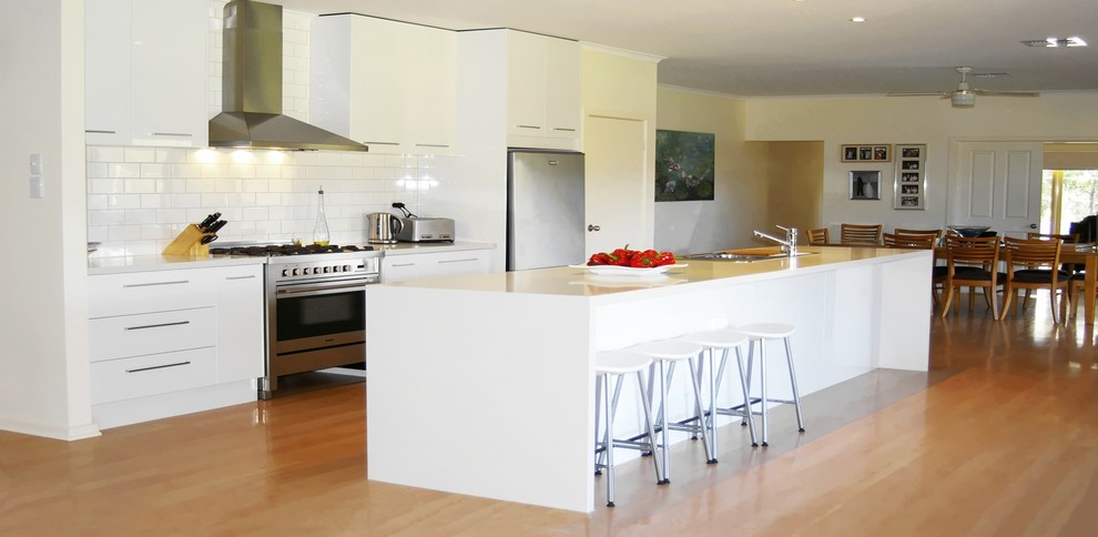 Mid-sized contemporary galley open plan kitchen in Adelaide with a double-bowl sink, flat-panel cabinets, white cabinets, quartz benchtops, white splashback, ceramic splashback, stainless steel appliances, medium hardwood floors, with island and white benchtop.