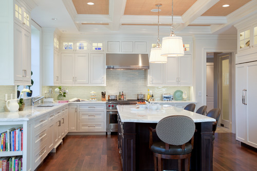 Light-filled transitional kitchen with white shaker RTA cabinets, lit glass transom uppers, pale subway-tile backsplash, stainless range hood, marble countertops, and a contrasting dark wood island with drum pendants.