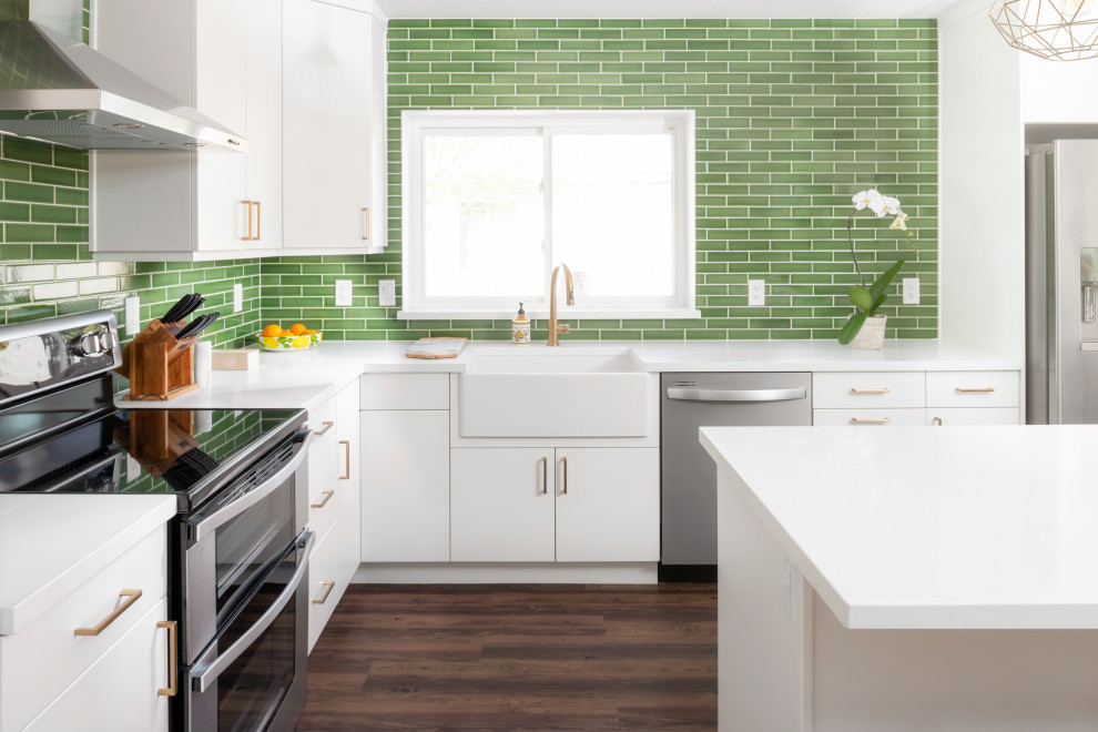 This is an example of a mid-sized midcentury l-shaped open plan kitchen in Phoenix with a farmhouse sink, flat-panel cabinets, white cabinets, quartz benchtops, green splashback, ceramic splashback, stainless steel appliances, vinyl floors, with island, brown floor and white benchtop.