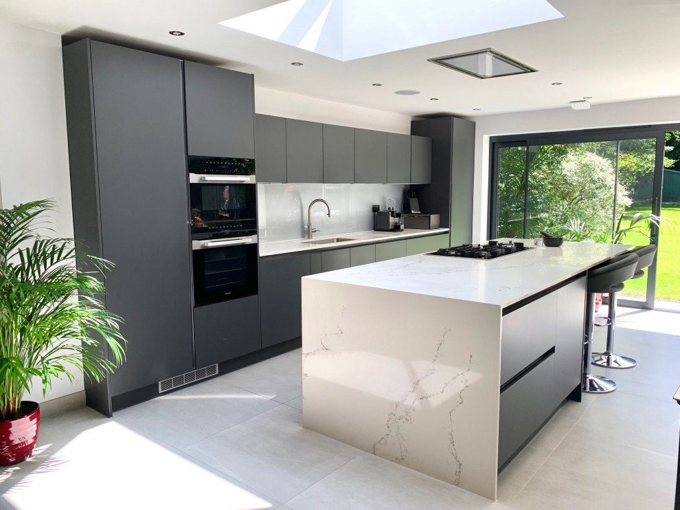 This is an example of a large contemporary single-wall open plan kitchen in Hertfordshire with a drop-in sink, flat-panel cabinets, black cabinets, marble benchtops, glass sheet splashback, black appliances, porcelain floors, with island, grey floor and white benchtop.