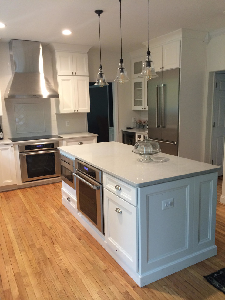 Open concept kitchen - mid-sized transitional l-shaped medium tone wood floor and brown floor open concept kitchen idea in DC Metro with a farmhouse sink, shaker cabinets, white cabinets, quartz countertops, white backsplash, subway tile backsplash, stainless steel appliances and an island