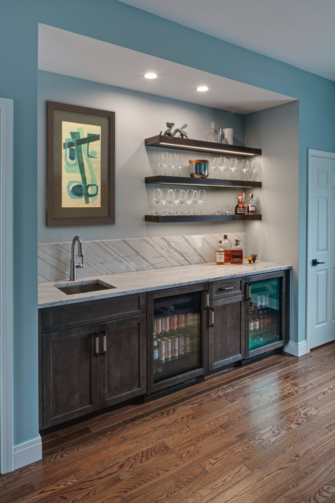 Mid-sized transitional single-wall wet bar in Chicago with an undermount sink, recessed-panel cabinets, brown cabinets, quartzite benchtops, grey splashback, stone slab splashback, medium hardwood floors, brown floor and grey benchtop.