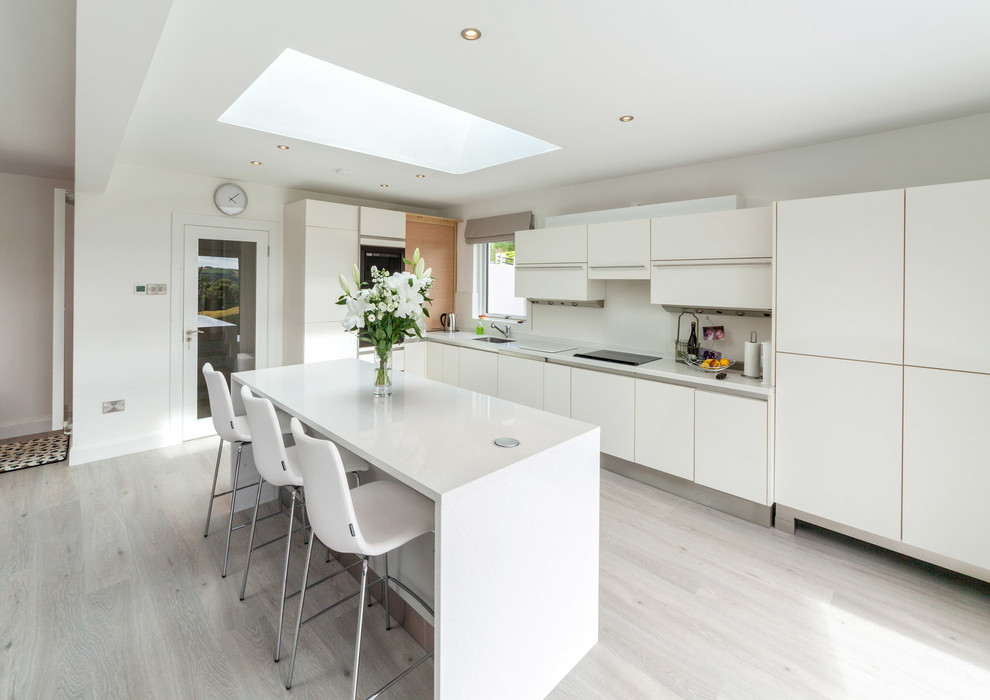This is an example of a mid-sized contemporary l-shaped open plan kitchen in Cork with a single-bowl sink, flat-panel cabinets, white cabinets, quartzite benchtops, white splashback, panelled appliances, linoleum floors and with island.