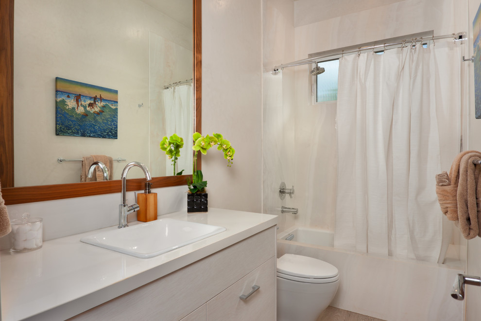 Photo of a mid-sized tropical 3/4 bathroom in Hawaii with porcelain floors, flat-panel cabinets, white cabinets, an alcove tub, a shower/bathtub combo, white tile, stone slab, white walls, a drop-in sink, limestone benchtops and a shower curtain.