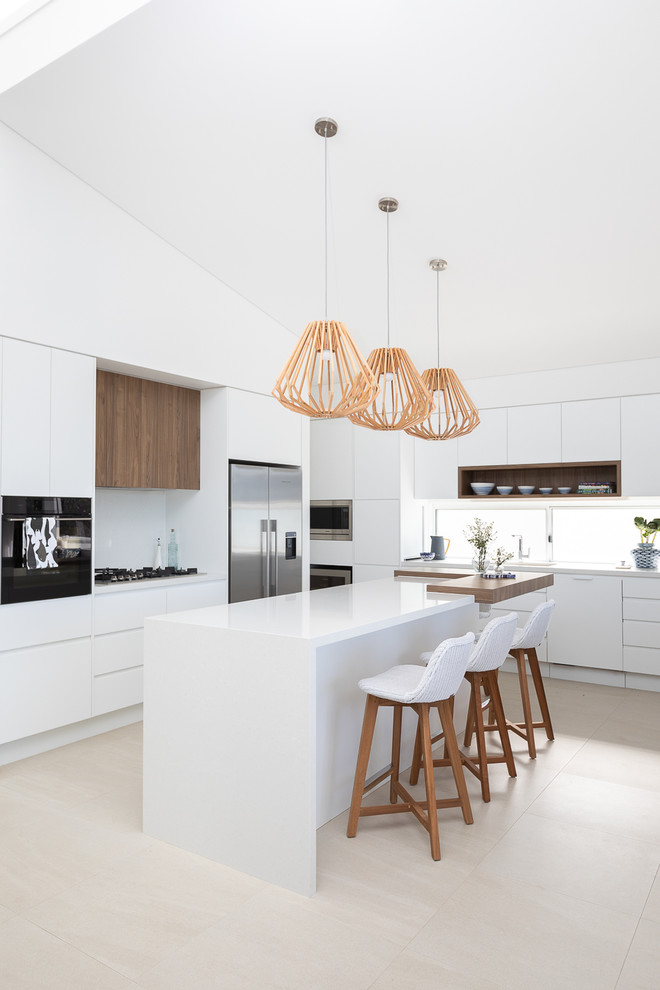 This is an example of a beach style l-shaped kitchen in Other with flat-panel cabinets, white cabinets, white splashback, stainless steel appliances, with island, beige floor and white benchtop.