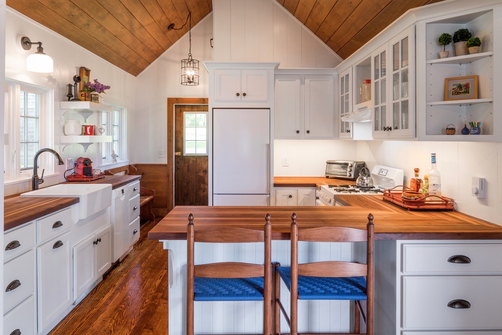 Inspiration for a beach style u-shaped kitchen in Portland Maine with a farmhouse sink, recessed-panel cabinets, white cabinets, wood benchtops, white splashback, white appliances, medium hardwood floors, a peninsula and brown floor.