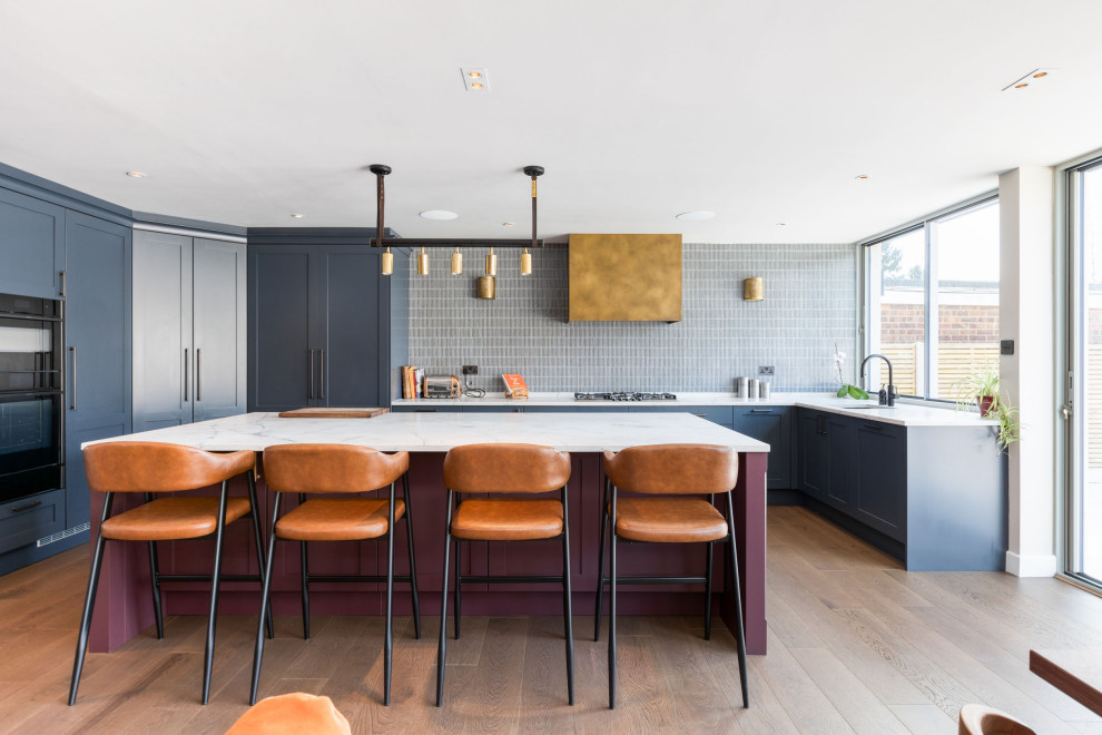 Expansive contemporary u-shaped open plan kitchen in Hertfordshire with shaker cabinets, blue cabinets, engineered stone countertops, blue splashback, black appliances, medium hardwood flooring, an island, brown floors, white worktops, a feature wall and a submerged sink.
