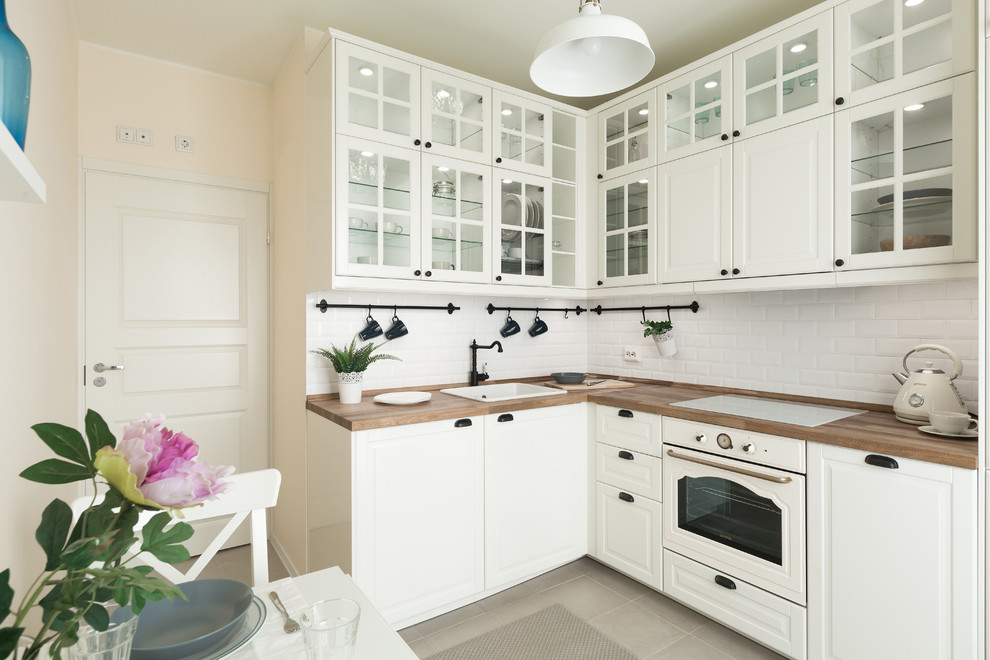 Design ideas for a transitional l-shaped eat-in kitchen in Saint Petersburg with a drop-in sink, glass-front cabinets, white cabinets, wood benchtops, white splashback, subway tile splashback, white appliances, no island and grey floor.