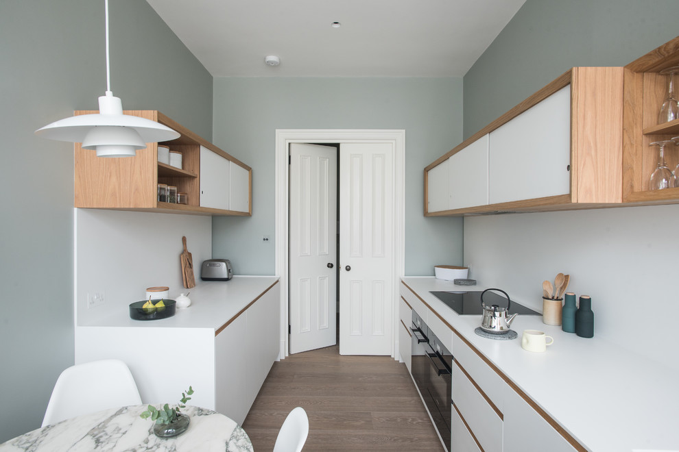 Design ideas for a small contemporary galley eat-in kitchen in London with an integrated sink, white cabinets, solid surface benchtops, white splashback, black appliances, medium hardwood floors, white benchtop, flat-panel cabinets and no island.