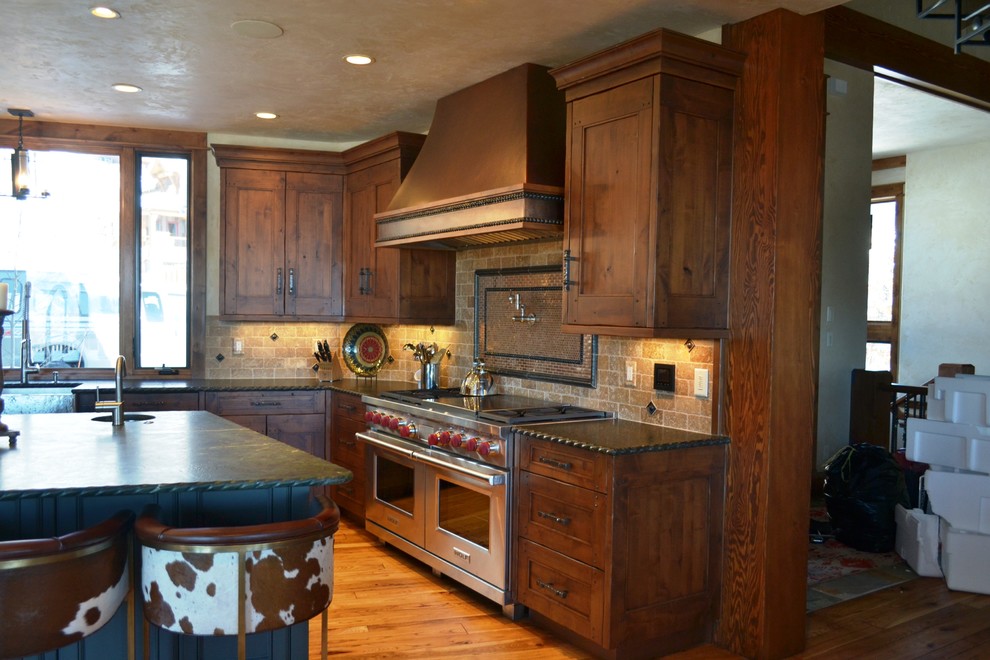 Example of a large mountain style u-shaped light wood floor and brown floor enclosed kitchen design in Denver with a farmhouse sink, recessed-panel cabinets, medium tone wood cabinets, granite countertops, beige backsplash, stone tile backsplash, stainless steel appliances and an island