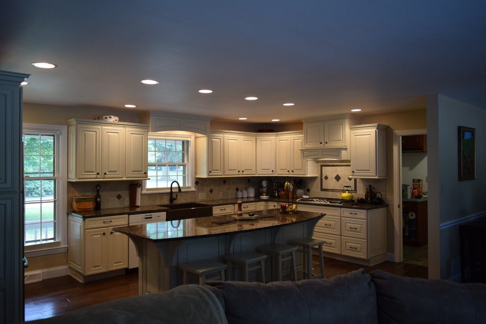 Transitional medium tone wood floor and brown floor kitchen photo with a farmhouse sink, raised-panel cabinets, white cabinets, quartz countertops, beige backsplash, ceramic backsplash, black appliances and an island
